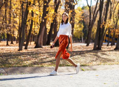 Sonbahar parkında yürüyen turuncu etekli esmer bir kadının vücut portresi.