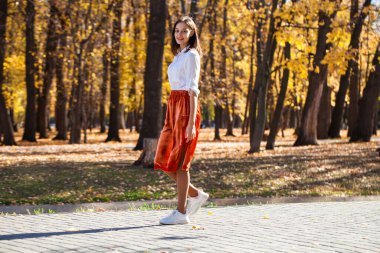 Sonbahar parkında yürüyen turuncu etekli esmer bir kadının vücut portresi.