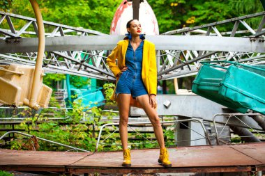 Bir lunaparkta atlıkarınca arka planında poz veren sarı ceketli ve kot pantolonlu manken.