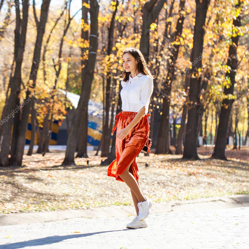 Full body portrait of a young brunette woman in orange skirt walking in autumn park
