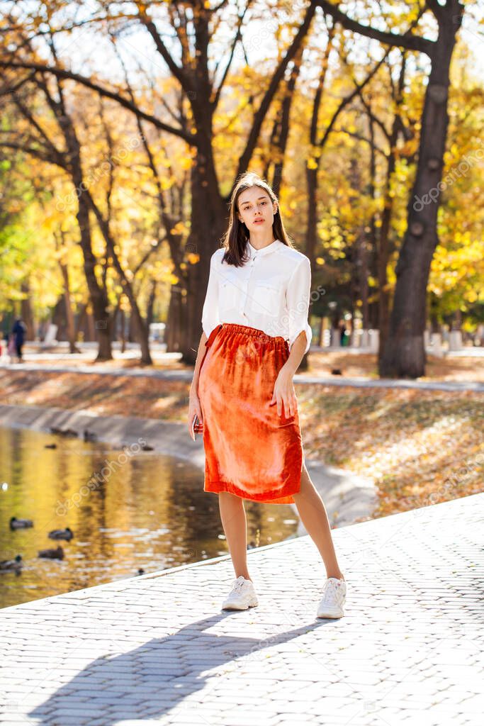 Full body portrait of a young brunette woman in orange skirt walking in autumn park