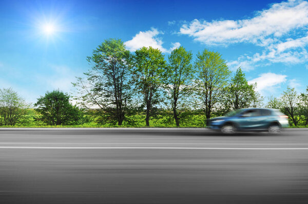 A blue crossover car driving fast on the countryside asphalt road in motion with green trees against blue sky with sun