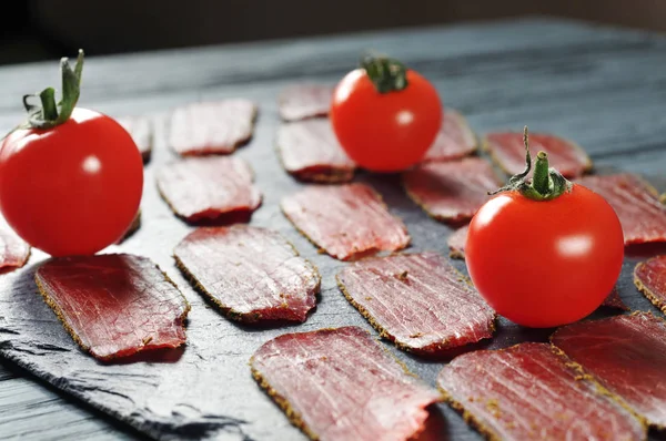 红色熏肉切片与香料和成熟的西红柿在一个黑色木板与木蓝表 — 图库照片