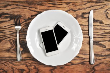 İki plaka üzerinde büyük siyah ekranlı akıllı telefonlar ile gümüş bıçak ve çatal koyu ahşap arka plan üzerinde beyaz