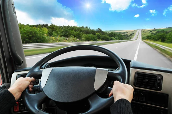 Truck dashboard with driver\'s hands on the steering wheel on the countryside road against blue sky with sun