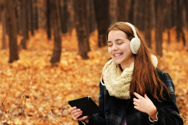 年轻美丽的女孩与温暖的围巾和耳罩持有黑色片剂在秋季森林中 背景模糊 — 图库照片