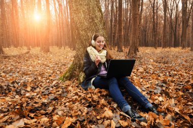 Genç kız beyaz kaşkollu ve düşen yapraklar ve güneş ışığı ile sonbahar ormandaki siyah dizüstü kullanarak kulaklık