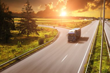 Blue truck with trailer on countryside road in motion with fields and green trees against sky with sunset clipart