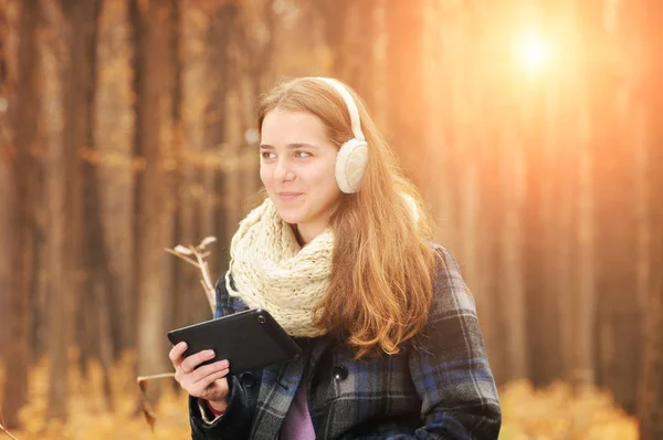 年轻的女孩与温暖的围巾和耳罩拿着黑色平板电脑在秋天森林以模糊的背景 — 图库照片