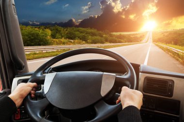 Truck dashboard with driver hands on steering wheel on countryside road against sky with sunset