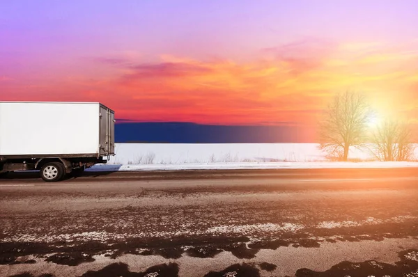 White box truck on countryside winter road with snow against sky with sunset