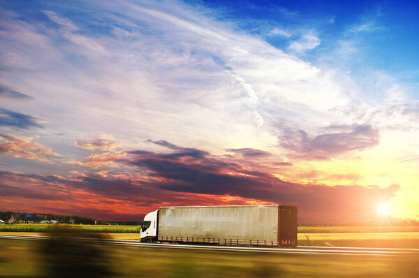 White truck with the trailer on the countryside road in motion with fields and green trees against night sky with sunset