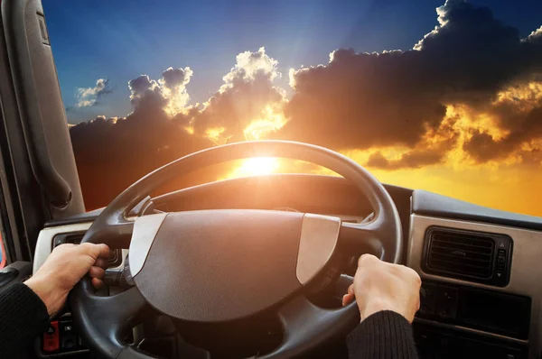 Truck dashboard with driver hands on steering wheel against sky with sunset