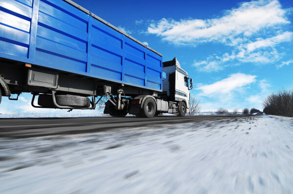White truck and blue trailer driving fast on winter countryside road with snow against blue sky with clouds