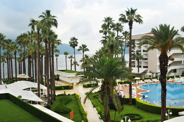 Hotel with green park, palm trees, pool and sea with mountains in clouds