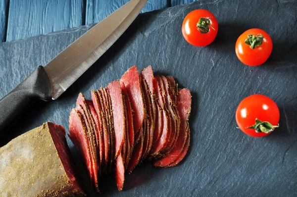 在深色切菜板和木制蓝色桌子上切碎的烤肉 黑色大刀和红色成熟的西红柿 — 图库照片