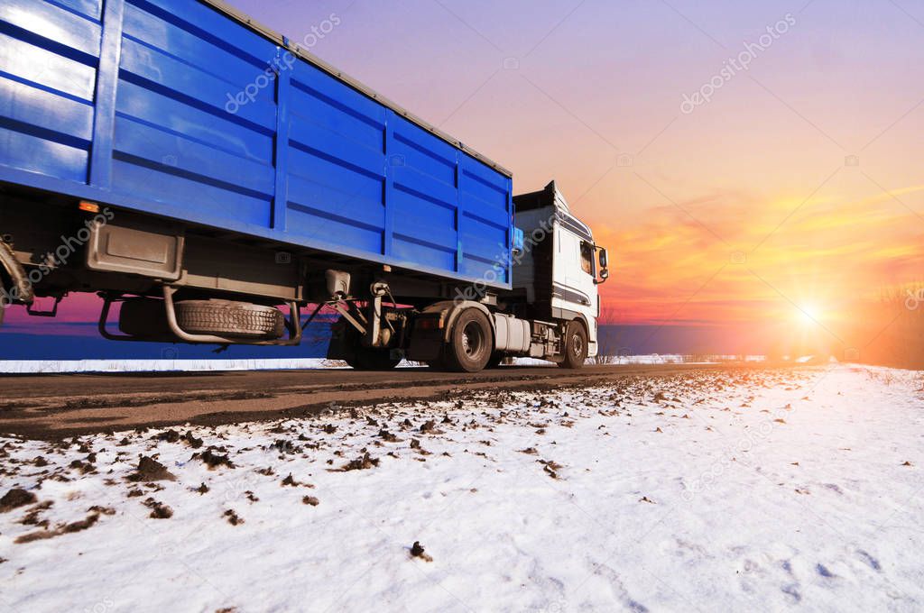 White truck and blue trailer driving fast on winter countryside road with snow against sky with sunset