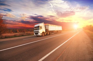 Big trucks and white trailers and cars on countryside road against sky with sunset clipart