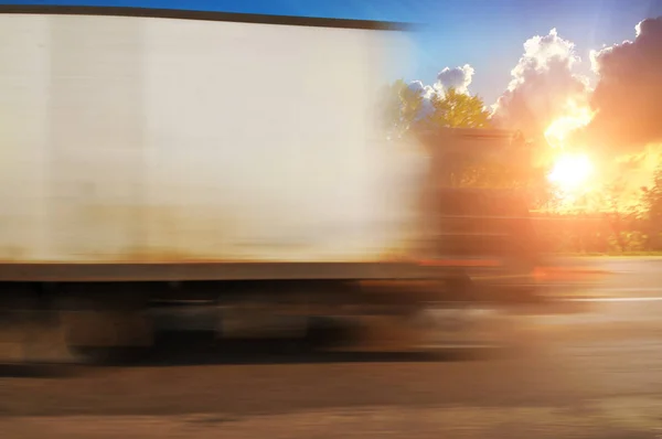 White big boxed truck driving fast on evening countryside road in motion with green trees and bushes against sky with sunset