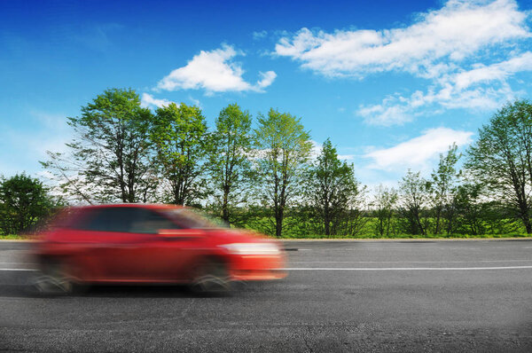 Red hatchback car driving fast on countryside road with green trees and bushes against blue sky with clouds