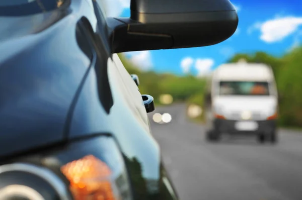 Black car side mirror and blurred background with cars on countryside road with green trees and blue sky with clouds