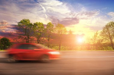 Kırmızı hatchback araba gün batımı ile gökyüzüne karşı yeşil ağaçlar ve çalılar ile hareket kırsal yolda hızlı sürüş