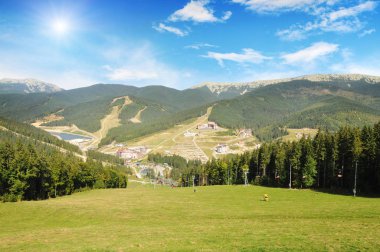 Yeşil orman, kayak merkezi binaları, telesiyejler ve yaz aylarında bulutlar ve güneş ile mavi gökyüzü ile Karpat dağlar 