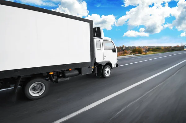 White big box truck with space for text driving fast on countryside road with trees and bushes against blue sky with clouds