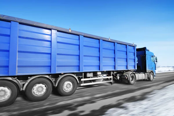 Blue truck and trailer driving fast on winter countryside road with snow against clear blue sky
