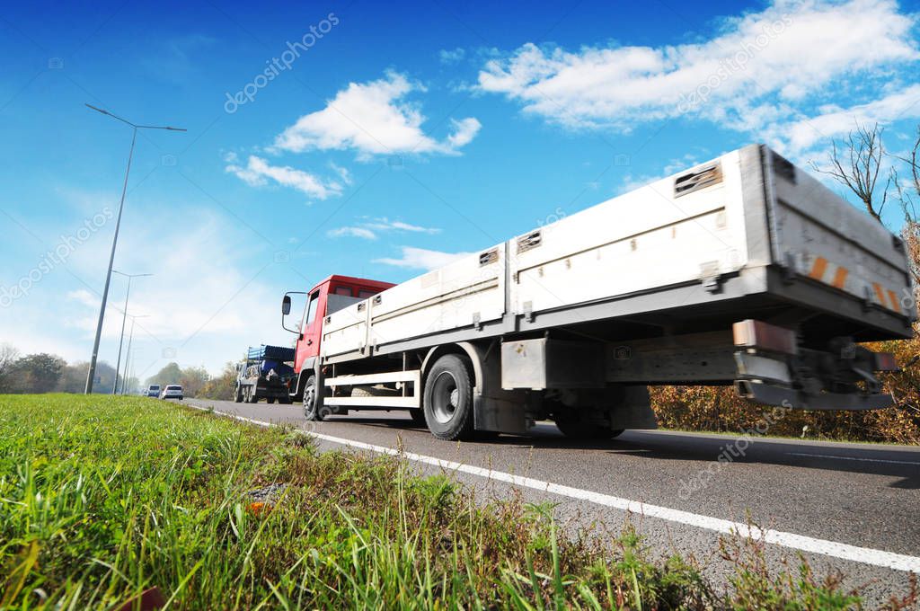 Flatbed truck and cars on countryside road against sky with clouds