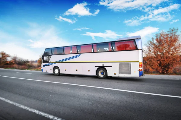 Big white bus on countryside road with trees and bushes against blue sky with clouds