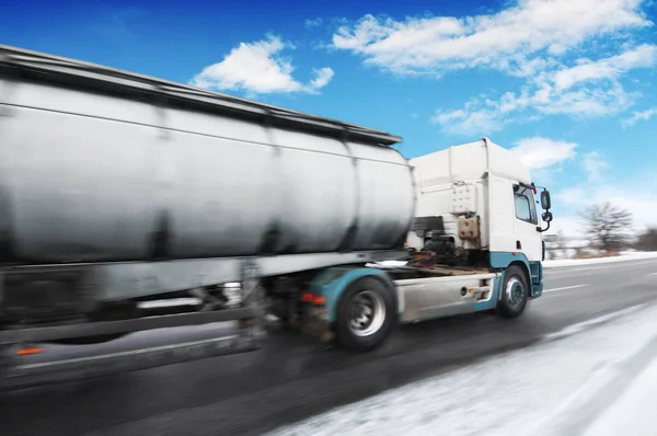 White big metal fuel tanker truck in motion shipping fuel on winter countryside road with snow against blue sky with clouds