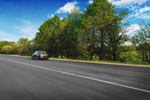 Black sedan car driving fast on countryside asphalt road with green trees against blue sky with clouds