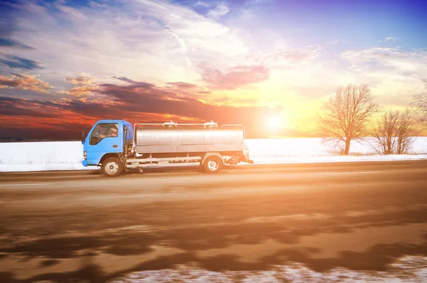Small blue truck shipping milk on countryside winter road in motion with snow against sky with sunset