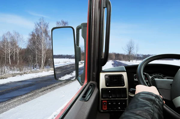 Truck dashboard with driver hand on steering wheel and side rear view mirror against winter road with white snow and blue sky