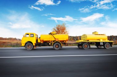 Yellow old soviet fuel tanker truck shipping fuel on the countryside road in motion with forest against blue sky with clouds clipart
