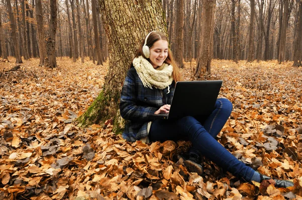 白いスカーフと耳フの若い女性は 落ち葉と秋の森の中で黒いノートパソコンを使用して — ストック写真