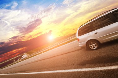Gökyüzüne karşı asfalt yolda gün batımlı gümüş bir minivan.