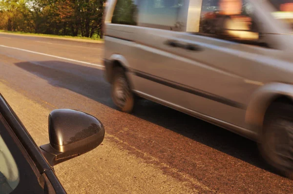 Siyah araba yan aynası ve diğer araba akşamları yeşil ağaçlarla kırsal yolda hızlı gidiyor.