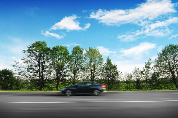 Black sedan car driving fast on the countryside asphalt road in motion with green trees against blue sky with clouds