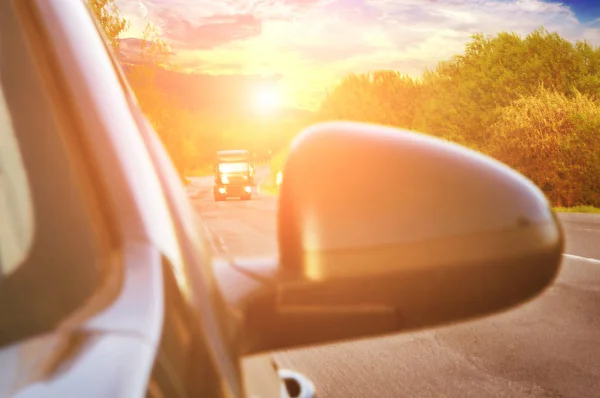 Blurred  black car side mirror and background with cars on the countryside road with green trees and sky with sunset