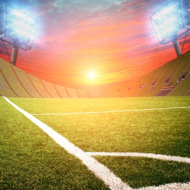 Futbol stadyumunun köşesinde yeşil çimenler ve parlak günbatımına karşı parlayan ışıklar.