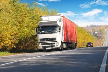 Sonbahar kırsal yollarında kırmızı römorklu ve arabalı bir kamyon Ağaçlar ve çalılar mavi gökyüzüne karşı bulutlu