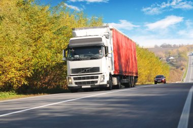 Sonbahar kırsalında kırmızı römorklu ve arabalı kamyon hareket halinde ağaçlar ve çalılar mavi gökyüzüne karşı bulutlarla