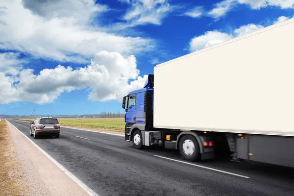 Big blue truck and white trailer with space for text and crossover car on countryside road against blue sky with clouds