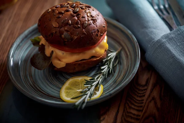 Dark burger with grain bread on dark ceramic plate, salad, rosemary and lemon