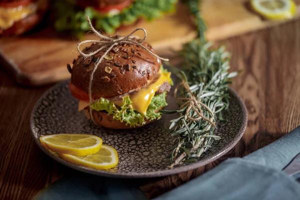 Dark burger with grain bread on dark ceramic plate, salad, rosemary and lemon
