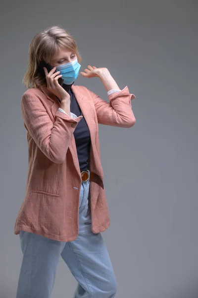 Rubia joven hembra en máscara protectora contra virus, teléfono inteligente hablando. — Foto de Stock