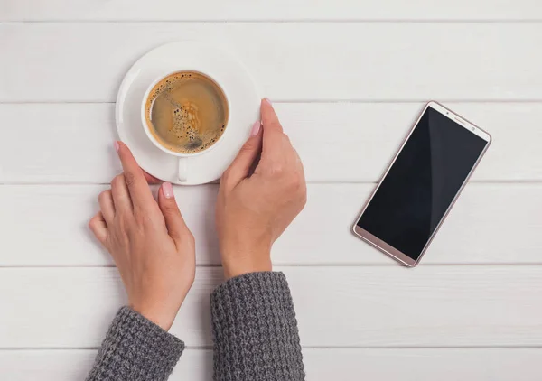 Γυναικεία χέρια σε γκρι πλεκτό πουλόβερ, κρατώντας ένα φλιτζάνι καφέ και το smartphone που βρίσκεται κοντά στο τραπέζι — Φωτογραφία Αρχείου