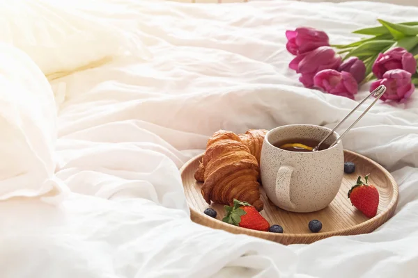 Uma xícara de chá e croissants na bandeja de madeira em pé na cama com lençóis brancos . — Fotografia de Stock